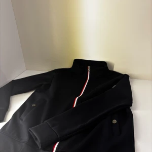 Svart bomberjacka från Moncler - Svart bomberjacka från Moncler med klassisk ståkrage och hel dragkedja i vitt och rött. Jackan har två fickor med tryckknappar och Moncler-logga på bröstet. Materialet är mjukt och känns sportigt, perfekt för en clean och stilren look. Detta är dress båda ingår 