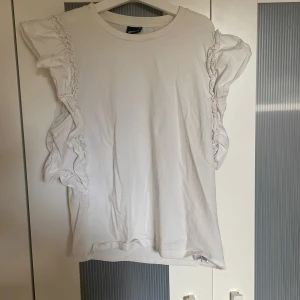 Vit volangtopp från Gina Tricot - Säljer en vit topp från Gina Tricot i storlek M. Toppen har korta ärmar med snygga volangdetaljer längs axlarna som ger en cool och trendig look. Materialet är mjukt och känns skönt mot huden, perfekt för dig som gillar stilrena plagg med lite extra edge.