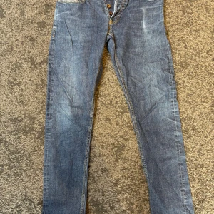 NN07 jeans  - Säljer ett par NN07 jeans i blå tvätt med snygga slitningar. Modellen har rak passform, normal midja och fem fickor. Jeansen är tillverkade i robust denim med gul kontrastsöm och knappgylf. Perfekt för dig som gillar tidlös stil.