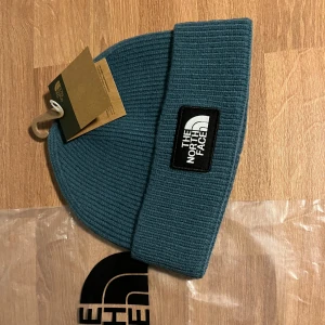 Blå  mössa från The North Face - Snygg blå/grön ribbstickad mössa från The North Face med uppvikt kant och tydlig logopatch framtill. Perfekt för kalla dagar och enkel att matcha med din jacka. Materialet är mjukt och värmande, och färgen är en djup blå ton.