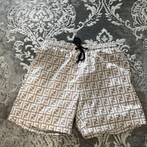 Beige Fendi shorts med FF-mönster - Snygga beige shorts från Fendi med ikoniskt FF-logomönster över hela tyget. De har svart snörning i midjan och är tillverkade i ett lätt syntetmaterial med meshfoder. Perfekta för sommaren och har en loose passform som ger skön vibe.