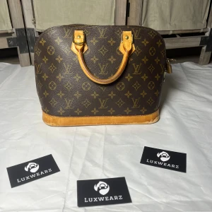 Louis Vuitton Alma - Snygg handväska från Louis Vuitton i klassiskt monogrammönster. Väskan är brun med ljusbruna detaljer och har rundade former, två handtag och dragkedja upptill. Tillverkad i skinn och canvas med ikoniska LV-loggor över hela ytan.