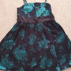 Klänning - Supersöt kortklänning från KappAhl med smala axelband och svart botten med grönt blommönster. Klänningen har markerad midja med satinliknande band och lätt utställd kjol. Perfekt för dig som gillar färg och mönster!