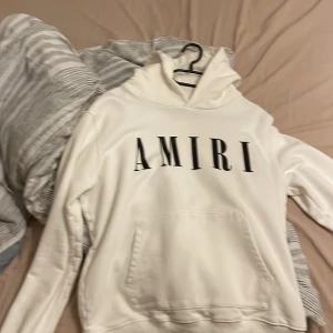 Vit hoodie från Amiri med logga. M/S - Säljer en clean vit hoodie från Amiri med stor svart logga framtill. Klassisk känguruficka, huva och snygga detaljer som metallbricka med Amiri-gravyr. Perfekt för dig som gillar stilrena streetwear-plagg.