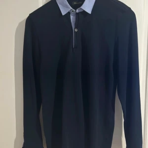 Mörkblå henley från Massimo Dutti - Snygg mörkblå henley från Massimo Dutti med ljusblå krage och knappslå. Skjortan har lång ärm och en clean, stilren look som passar perfekt till jeans eller chinos. Casual fit för en avslappnad vibe.