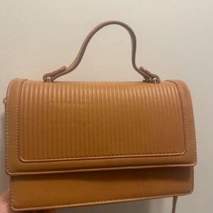 Beige handväska med ribbad framsida - Snygg beige handväska i skinn med ribbad struktur på framsidan och praktiskt handtag. Insidan har flera fack och dragkedjor, fodrad i rött. Perfekt för dig som vill ha en stilren och trendig väska med smart förvaring.