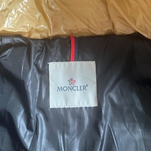 Beige pufferjacka med päls från Moncler - Snygg vintage beige pufferjacka från Moncler med glansig finish och huva med pälsdetalj. Jackan har dragkedja framtill och är quiltad för extra värme. Perfekt för kalla dagar och ger en riktigt clean look. Moncler logon på armen har trillat av.