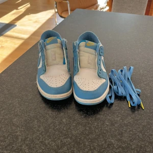 Nike Dunk Low blå och vita sneakers - Nike Dunk Low sneakers i fräsch blå och vit färg med klassisk design. Skorna har perforerad tå, rund tå och platt sula. Medföljer matchande blå skosnören med gula detaljer. Ovandel i skinn och textil, ikonisk Nike-logga på sidan och tungan.