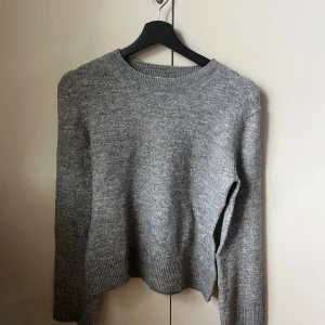 Grå stickad tröja med rund hals - Mysig grå stickad tröja med rund hals och ribbade muddar vid ärmslut och nederkant. Perfekt för kyliga dagar och enkel att matcha med jeans eller kjol. Långärmad och har en klassisk, normal passform som funkar till många olika stilar.