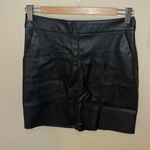 Svart skinnkjol från Vero Moda - Snygg svart skinnkjol från Vero Moda i storlek XS. Kjolen har en rak passform och en cool silverfärgad dragkedja med ringdetalj bak. Insidan av kjolen har ett mjukt material som gör den väldigt bekväm att ha på sig. Knappt använd så inga defekter, endast lite skrynklig vilket går att fixa!🥰
