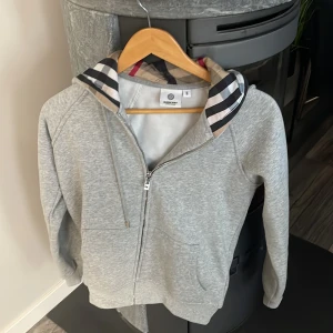 Grå hoodie från Burberry med dragkedja - Snygg grå hoodie från Burberry med klassiskt rutigt foder i huvan och dragkedja framtill. Tröjan har långa ärmar, snörning i huvan och två fickor. Perfekt för dig som gillar stilrena och exklusiva plagg med en twist.. passar nån som är mellan ca 165-175. Oanvänd 