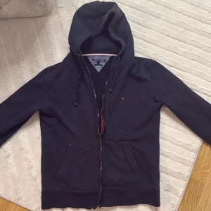 Svart hoodie från Tommy Hilfiger - Svart hoodie från Tommy Hilfiger med dragkedja, huva med snörning och två fickor framtill. Klassisk liten logga broderad på bröstet och vintage fit-passform. Perfekt för chill dagar och snygg streetstyle.