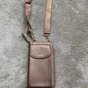 Brun axelväska från Ulrika - Snygg brun axelväska från Ulrika i skinnimitation med justerbar rem. Väskan har flera fack, dragkedja och plats för kort. Perfekt storlek för mobilen och det viktigaste. Stilren design med diskret logga på framsidan.