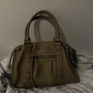 Snygg city väska  - Snygg beige/khaki färgad handväska/axelväska i  skinn med dubbla handtag och snygga metalldetaljer. Väskan har  dragkedja upptill, litet ytterfack med dragkedja och dekorativa spännen framtill. Det finns stort utrymme inuti med två rymliga fack. Väskan har användes och kan ha lite svanker och tecken på användning men fyller fortfarande dess funktion. Och även tvättat den så gott jag kan. Finns en skavank på ena axel bandet. Nypris runt 1500kr. (Kan skicka fler bilder