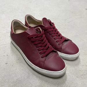 Axel Arigato Sneakers - Snygga vinröda sneakers från Axel Arigato i skinn med matchande vinröda snören och vit platt sula. Diskret guldtext på sidan och mockadetalj vid hälen. Perfekta för dig som gillar stilrena och trendiga skor med premiumkänsla.