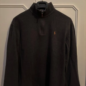 Mörkgrå half zip tröja Polo Ralph Lauren - Mörkgrå långärmad tröja från Polo Ralph Lauren med klassisk broderad logga på bröstet. Tröjan har en hög krage med half zip-dragkedja och är tillverkad i mjuk bomullsmix. Perfekt för lager-på-lager och chill stil.