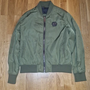 Olivgrön bomberjacka från Plein - Snygg olivgrön bomberjacka från Plein med broderad döskalle och stjärnor på ryggen. Jackan har ribbade muddar, dragkedja framtill och två fickor. Tillverkad i glansig polyester med klassisk bomberkrage och lång ärm. Perfekt jacka nu under tidig höst. OBS postas ej bara direkta möten