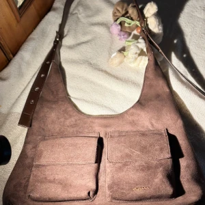Brun mocka axelväska med fickor - Snygg brun axelväska i mocka med två stora framfickor som stängs med magnetknapp. Väskan har justerbar axelrem i mörkbrunt skinn och en mjuk, slät yta. Perfekt för dig som gillar praktiska detaljer och stilren design.