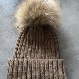 Brun ribbstickad mössa med pälsboll - Snygg brun ribbstickad mössa med stor fluffig pälsboll på toppen. Tror den är från Chelsea, och den är även oanvänd. Äkta päls 