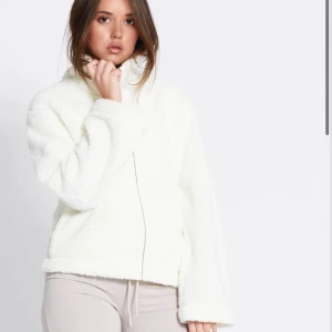 Vit teddyjacka med dragkedja - Mysig vit teddyjacka med hög krage och dragkedja framtill. Jackan har en loose passform och är tillverkad i fluffigt teddy-material som ger en riktigt skön och trendig look. Perfekt för kyliga dagar när du vill vara både varm och snygg.