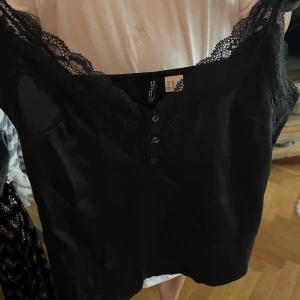 Svart spetslinne - Säljer ett svart linne från H&M divided i storlek XS. Linnet har v-ringning med spetskant och tre små knappar framtill. Materialet är glansigt och känns mjukt mot huden, perfekt för att lyfta vilken outfit som helst.