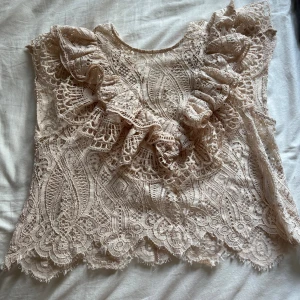 Beige spetsblus med volang från Zara - Supersöt beige spetsblus från Zara med volangdetaljer framtill och vackert mönstrad spets. Blusen är ärmlös och har en liten knapp i nacken. Perfekt för dig som gillar romantisk och feminin stil.