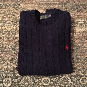 Mörkblå kabelstickad tröja Polo Ralph Lauren - Kabelstickad mörkblå tröja från Polo Ralph Lauren med rund halsringning och klassisk röd broderad logga på bröstet. Tröjan har långa ärmar och ribbade muddar. Perfekt för dig som gillar preppy och stilren look.