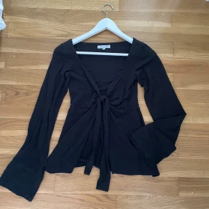 Svart omlottopp med knyt från Designbysi - Svart omlottopp från Designbysi med långa utsvängda ärmar och djup v-ringning. Toppen har knyt framtill som ger en snygg och trendig look. 