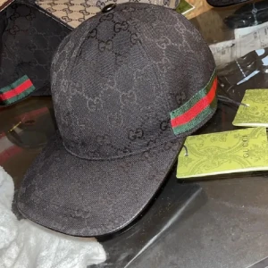 Svart Gucci keps med GG-mönster - Svart keps från Gucci med klassiskt GG-monogram över hela ytan och ikoniska gröna och röda ränder på sidan. Kepsen har böjd skärm och justerbar rem baktill. Materialet är slitstark bomull med insida i svart tyg. Perfekt för dig som vill ha en lyxig streetstil.