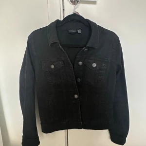 Svart jeansjacka  - Svart jeansjacka från Vero Moda med klassisk krage, knäppning framtill och två bröstfickor med knappar. Jackan har raka ärmar och en stilren, tidlös look som passar perfekt till alla outfits. Materialet är denim och jackan har en normal passform.