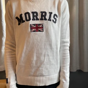 Vit stickad tröja från Morris - Vit stickad tröja från Morris med broderad logga och brittisk flagga på bröstet. Tröjan har ribbade muddar vid ärmslut och nederkant, samt rund halsringning. Snyggt och klassiskt college-inspirerat stuk som passar till jeans eller chinos.
