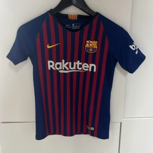 FC Barcelona matchtröja Nike M - Säljer en officiell FC Barcelona matchtröja från Nike med Dri-Fit material. Tröjan är blå med röda vertikala ränder, har klubbmärke på bröstet och Rakuten som sponsor. Kortärmad och med gula detaljer vid halsen. Perfekt för dig som älskar Barca!