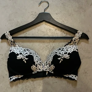 Svart intemissmi bh med spetsdetaljer - Snygg svart bh med vit broderad spets i blommigt mönster längs kupor, axelband och nederkant. Axelbanden är justerbara och materialet känns mjukt och följsamt mot huden. Perfekt för dig som gillar detaljer och vill ha något extra under kläderna. Skriv innan köp 