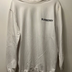 Vit sweatshirt från Burberry med logga - Vit sweatshirt från Burberry i mjuk bomull. Framsidan har en stilren Burberry-text i svart och baksidan pryds av ett stort TB-monogram i blått och vitt mönster. Tröjan har rund hals, långa ärmar och ribbade muddar. Perfekt för dig som gillar exklusiva streetwear-vibbar.