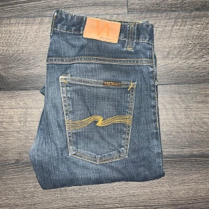 Nudie Jeans Thin Finn - Säljer ett par Nudie Jeans i modellen Thin Finn med klassiska orange sömmar och Nudie-logga på bakfickorna. Jeansen har smal passform och är tillverkade i stretchig bomull för en bekväm känsla. Midja 39cm, längd 104cm. JAG SKICKAR EJ FLER BILDER NÄR JAG BÄR JEANSEN! för storleksguide hänvisar jag till måtten och för bättre inblick i passformen rekommenderar jag att googla på modellnamnet :) s3