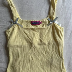 Gul ribbad linnetopp från Edikted  - Supersöt ljusgul ribbad linnetopp från Edikted med breda axelband och små silvriga rosetter vid axlarna. Toppen har en tight passform och är perfekt för varma dagar. Materialet är mjukt och stretchigt för extra komfort. Aldrig använt