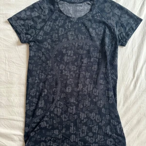 Svart leopardmönstrad träningstopp Lululemon - Superskön svart träningstopp från Lululemon med grått leopardmönster. T-shirten har rund halsringning, korta ärmar och figurnära passform. Perfekt för gymmet eller löprundan, tillverkad i ett stretchigt och ventilerande syntetmaterial. Har klippt av taggen på insidan.