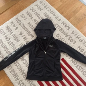 Svart EA7 hoodiejacka med dragkedja - Svart hoodiejacka från Emporio Armani EA7 med vit logga på bröstet och stort tryck längs ena ärmen. Jackan har hel dragkedja framtill, huva och är tillverkad i ett mjukt, sportigt polyestermaterial. Perfekt för en sportig och stilren look.