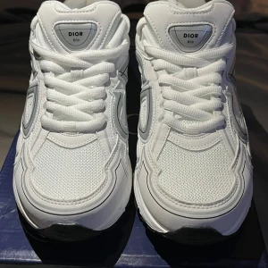 Dior B30 vita sneakers - Säljer ett par Dior B30 sneakers i helvitt med reflekterande silverdetaljer och stora CD-loggor på sidorna. Skorna har meshpaneler, skinnpartier och tjock vit sula. Klassisk snörning och sportig vibe, perfekt för dig som gillar exklusiva och trendiga sneakers. Storlek 43. Pris kan diskuteras.