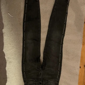 Svarta jeans med nitar längs benen - Snygga svarta jeans med raka ben och silverfärgade nitar längs sidorna. Jeansen har klassisk femficksdesign och är tillverkade i jeansmaterial. Perfekt för dig som vill ha en cool och edgy look med extra detaljer.