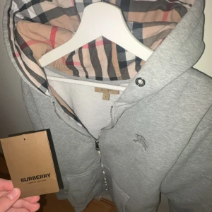 Grå Burberry kofta - Snygg grå hoodie/kofta från Burberry med klassiskt rutigt foder i huvan och broderad logga på bröstet. Tröjan har dragkedja framtill, två fickor och långa ärmar. Perfekt för dig som vill ha en stilren och lyxig look. Storlek L men sitter mer som M. (snörena är avtagna)