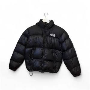 Svart pufferjacka från The North Face - Svart pufferjacka från The North Face med broderad logga på bröstet och ryggen. Jackan har hög krage, dragkedja framtill och justerbar dragsko nertill. Yttermaterialet är nylon och fodret är fyllt med dun för extra värme. Perfekt för kalla dagar.Har ett litet hål på insidan.