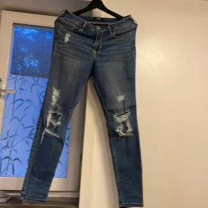Blå skinny jeans från Hollister - Snygga blå skinny jeans från Hollister med slitningar på knäna och bakfickan. Klassisk femficksmodell med dragkedja och knapp. Jeansen har en smal passform och är tillverkade i stretchigt denim för extra komfort.