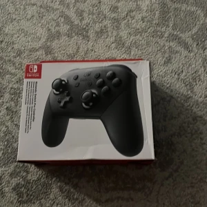 Nintendo Switch Pro Controller - Nintendo Switch Pro Controller i originalförpackning. Ergonomisk design för långa spelsessioner, trådlös anslutning och precis styrning. Kontrollen och kartongen är i gott skick, med mindre märken på kartongen. Perfekt för Switch-spel i TV- eller bords-läge.