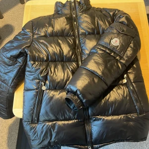  Moncler Maya  - Svart pufferjacka från Moncler med glansig finish, hög krage och flera dragkedjor. Jackan har Moncler-logga på ärmen och coolt serietidningsmärke på insidan. Perfekt för kalla dagar och har en riktigt snygg streetstil.