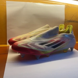Adidas F50 fotbollsskor vit/röd - Adidas F50 fotbollsskor med strömlinjeformad design i vitt, rött och neon-gult. Skorna har metall- och plastdobbar, stickad ovandel och svarta detaljer på sidan. Adidas-logga på hälen och F50-tryck på ovansidan. Perfekt för dig som vill ha snabba och lätta skor på planen.