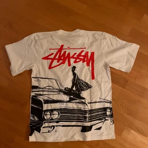 Vit Stüssy t-shirt med tryck - Vit t-shirt från Stüssy med rött logotryck fram och stort grafiskt tryck på ryggen med bil och siluett. Klassisk rund hals och korta ärmar. Skön streetwear-stil, perfekt för dig som gillar bold prints och ikoniska märken. Inte org