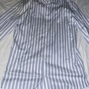 Randig Tommy Hilfiger skjorta - Klassisk skjorta med blå och vita vertikala ränder och knäppning framtill. Skjortan har lång ärm och krage, perfekt för en stilren look. Tillverkad i mjuk bomull som känns skön mot huden.