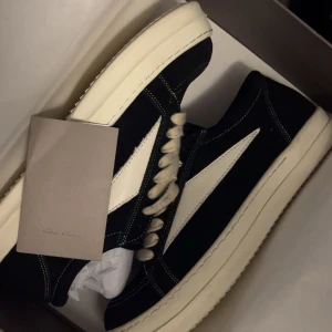 Rick Owens - Säljer ett par svarta Rick Owens sneakers med vit sula och vita detaljer på sidan. Skorna har snörning, kontrastsömmar och är tillverkade i mocka och skinn. Klassisk låg modell med chunky sula och stilren design, perfekt för dig som gillar streetwear och exklusiva märken.