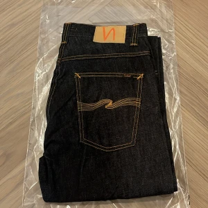 Helt nya Nudie jeans co - Slim Jim - W32 L32 - Helt nya och oanvända Nudie Jeans Co Slim Jim i den klassiska tvätten Dry Streaky – fortfarande i originalförpackning (se bilder).  Detta är en tidlös modell från Nudie Jeans med rak passform, perfekt balans mellan klassiskt snitt och modern stil, tillverkade i högkvalitativ japansk denim.  Detaljer: • Modell: Slim Jim • Storlek: W32 / L32 • Färg: Dry Streaky (mörk indigo med subtilt randig struktur i tyget) • Passform: Regular slim – normal midja, lätt avsmalnande ben • Material: 100% bomull